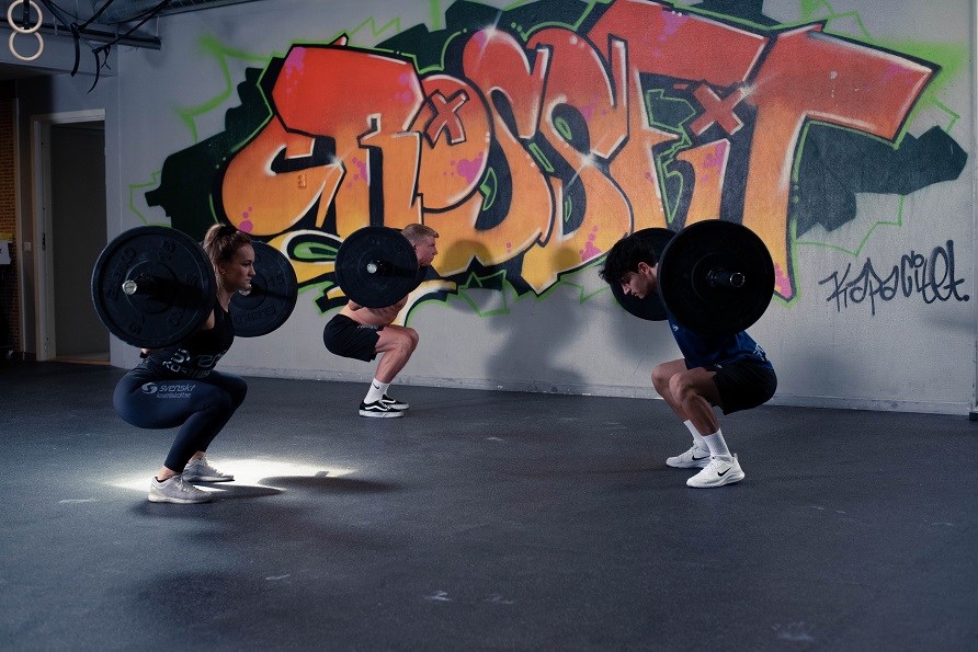 En kvinde og to mænd i et fitnesscenter laver squat med en stang på ryggen,